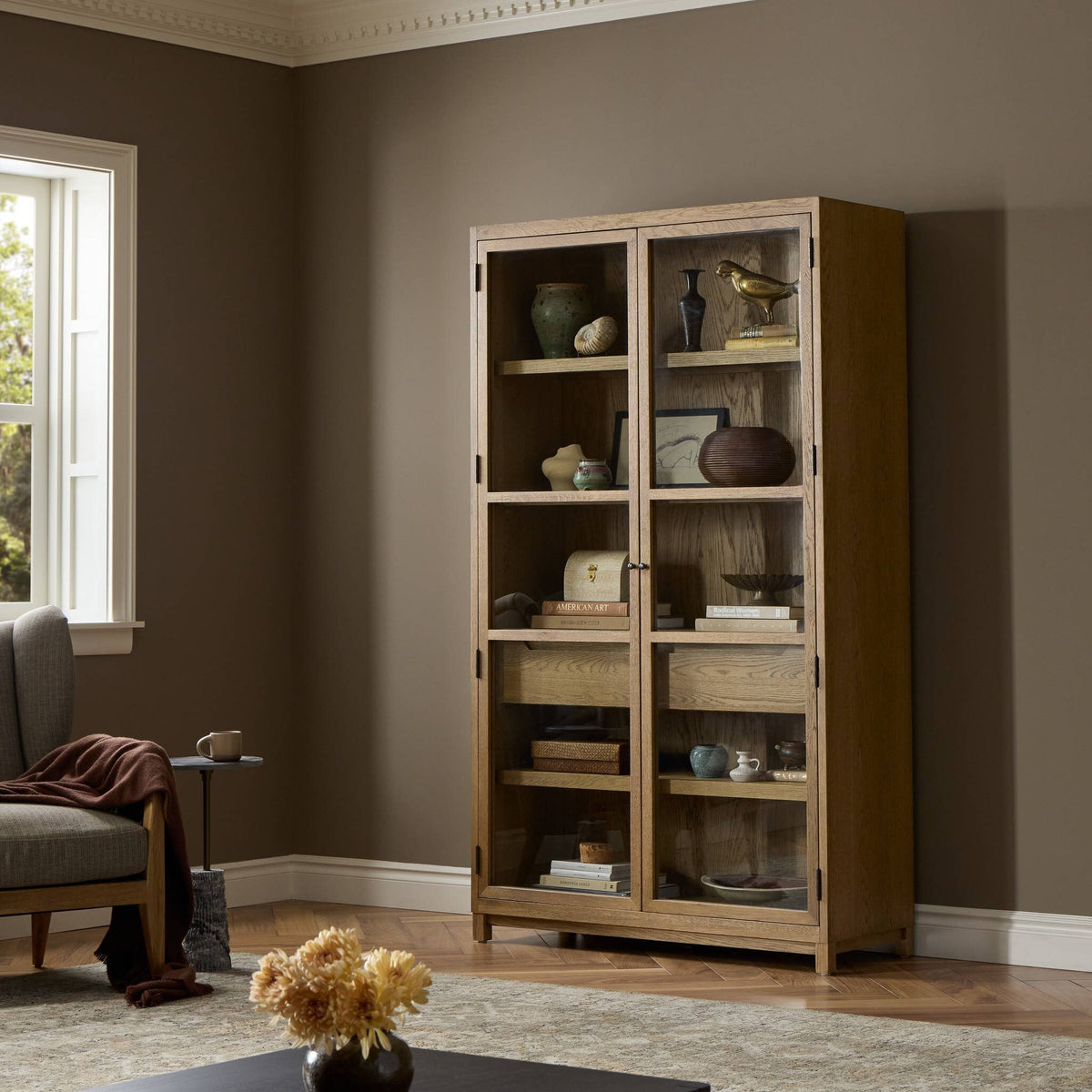 Kyra Cabinet in drifted oak with glass shelves showcasing decorative items.